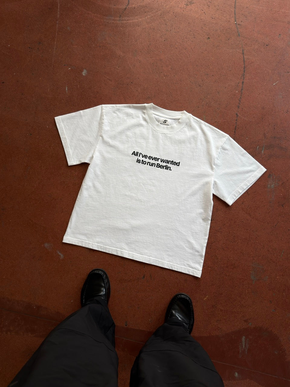 BERLIN TEE laid flat on a red concrete floor